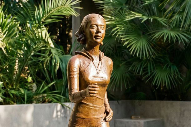 Statue en bronze représentant une femme en mouvement, installée en extérieur sur un patio entouré de feuillages tropicaux.